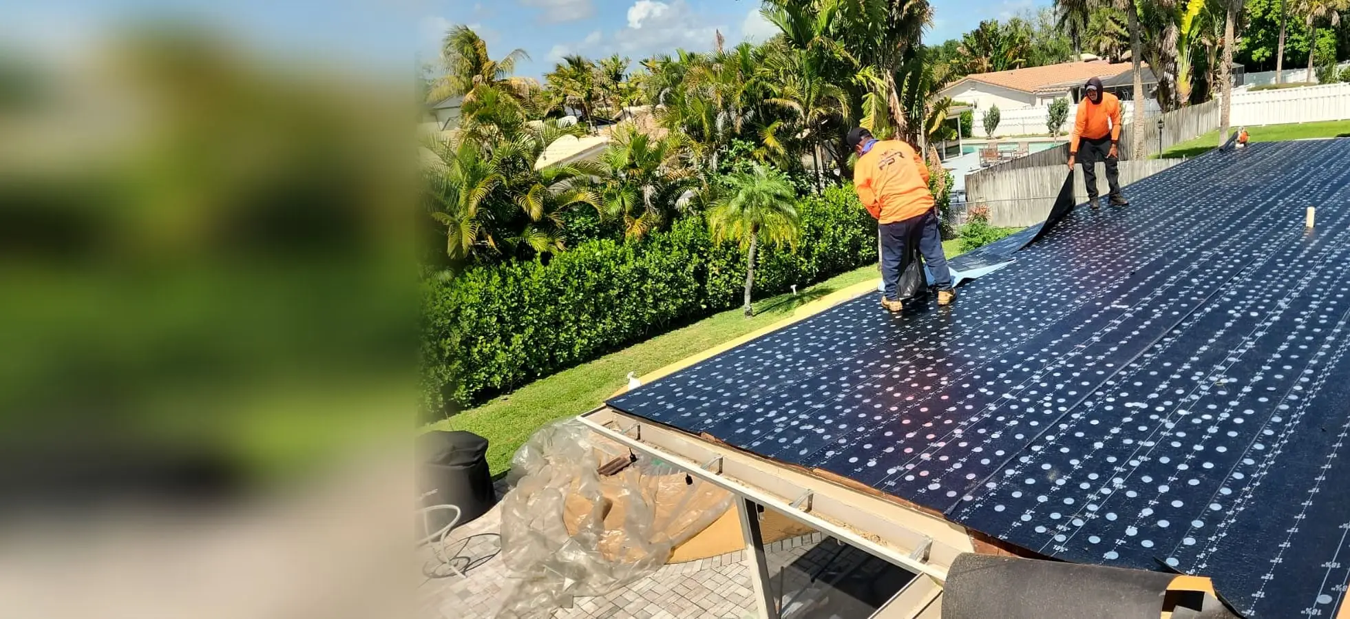 Two PSR crew members in orange shirts laying underlayment across a flat roof, with palm trees and a hedge behind