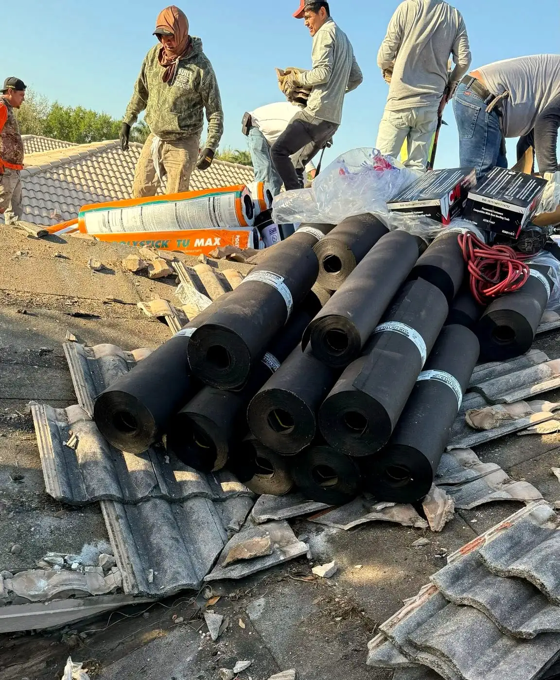 A crew tearing off old concrete tile, with rolls of underlayment staged on the roof